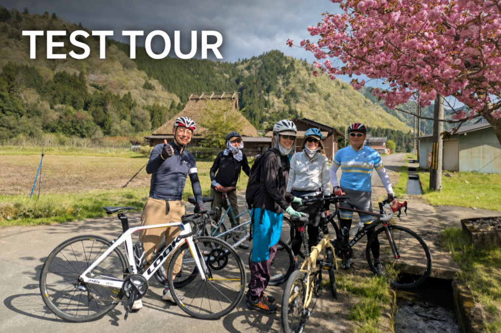 Cyclists posing with their bikes in a traditional village in Miyama, Kyoto, with cherry blossoms and thatched-roof houses in the background.